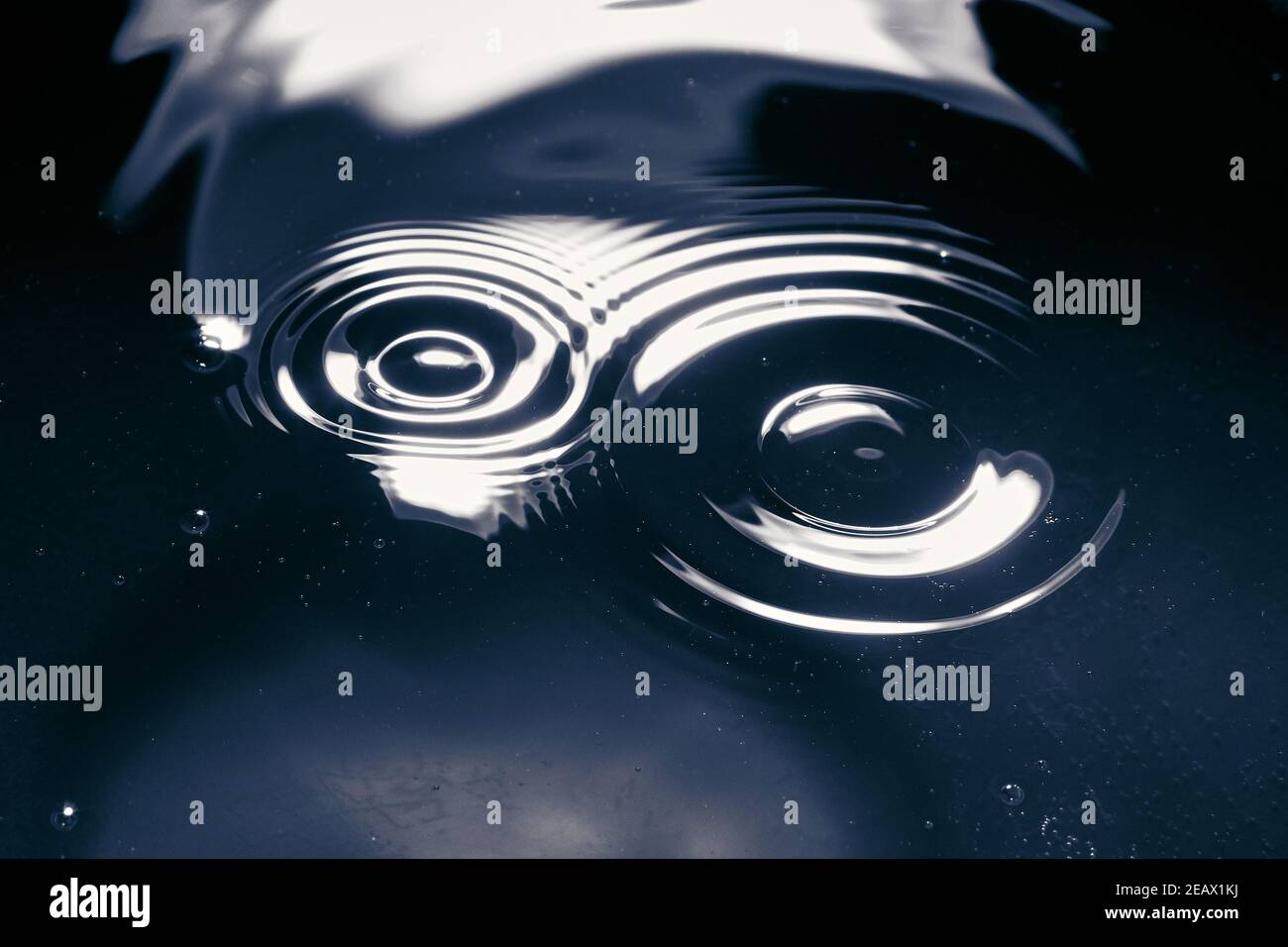 Wassertropfen fallen auf Wasser und verursachen Wellen in Kreisen auf blauem Hintergrund mit weißen Reflexen. Stockfoto