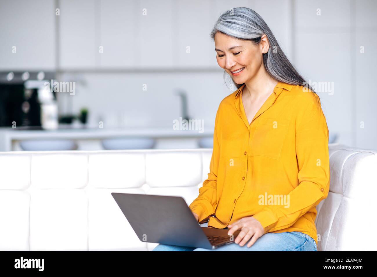 Entspannt mittleren Alters grauhaarige asiatische Frau in lässige stilvolle Kleidung gekleidet sitzt zu Hause im Wohnzimmer, mit Laptop, online kaufen, Arbeit von zu Hause aus, o Stockfoto