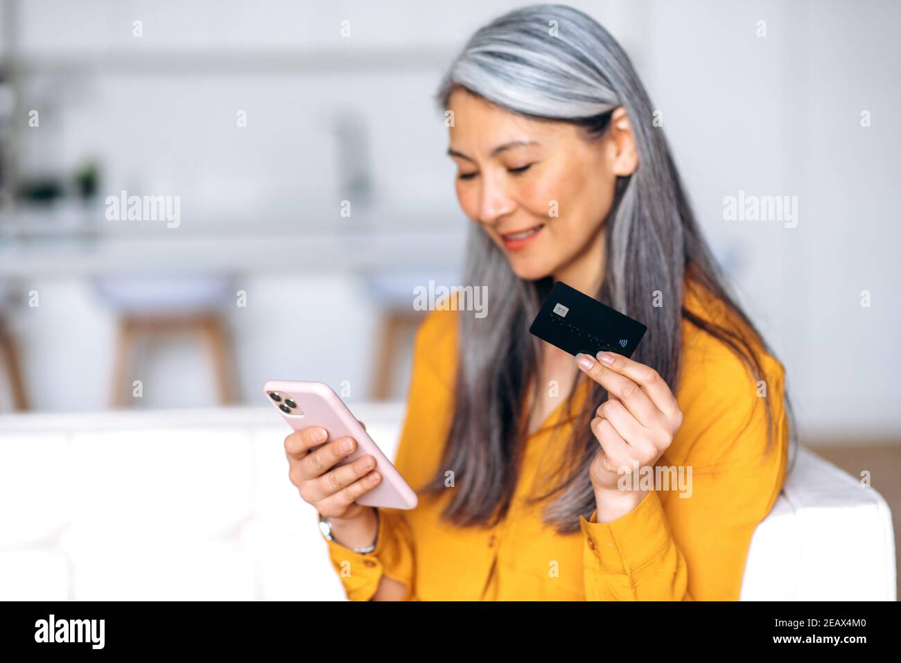 Defokussierte ältere grauhaarige Frau ist online einkaufen. Fröhliche asiatische grauhaarige Frau verwendet die Kreditkarte und Handy für den Einkauf und die Zahlung o Stockfoto