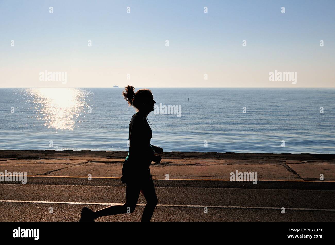 Chicago, Illinois, USA. Eine einsame Joggerin wird gegen die Reflexion der aufgehenden Sonne am Lake Michigan in der Nähe von North Avenue Beach silhouettiert. Stockfoto
