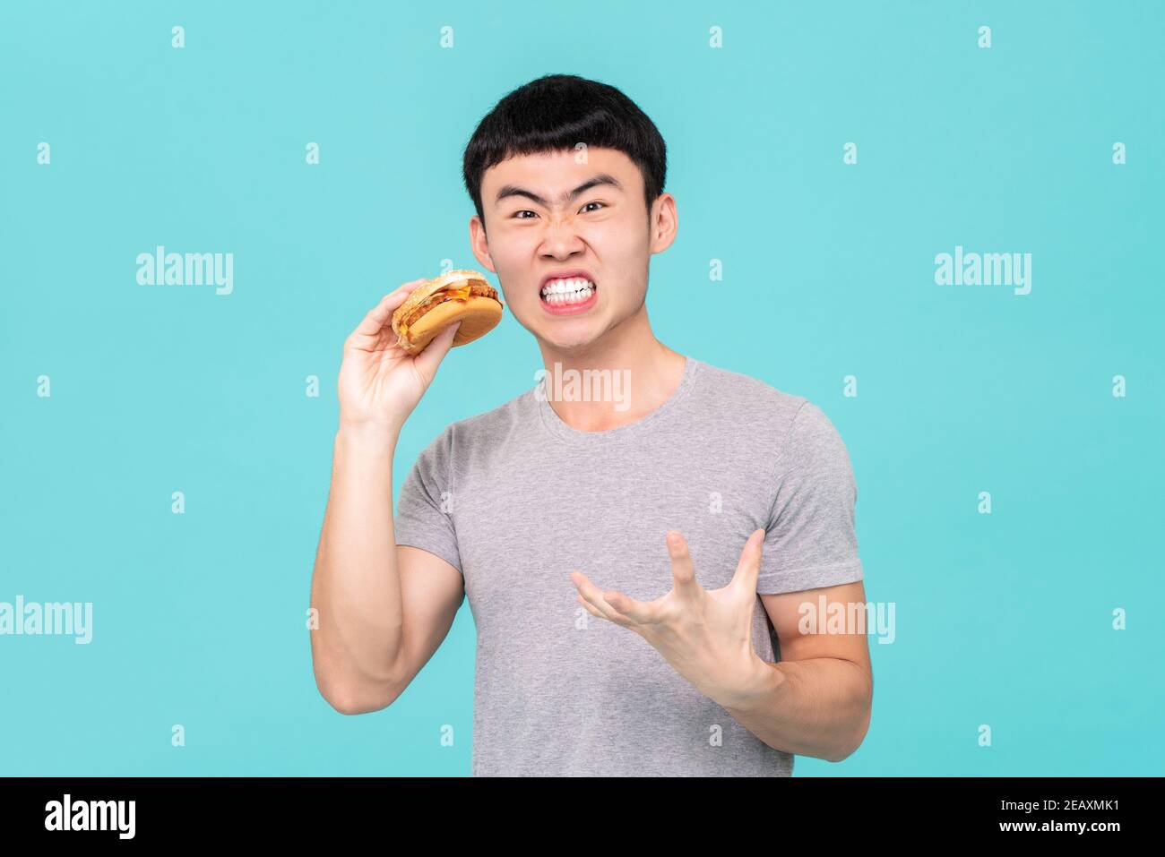 Diät hungrigen asiatischen Mann mit lustigen Gesichtsausdruck beim Halten hamburger in der Hand isoliert auf hellblauem Hintergrund Stockfoto