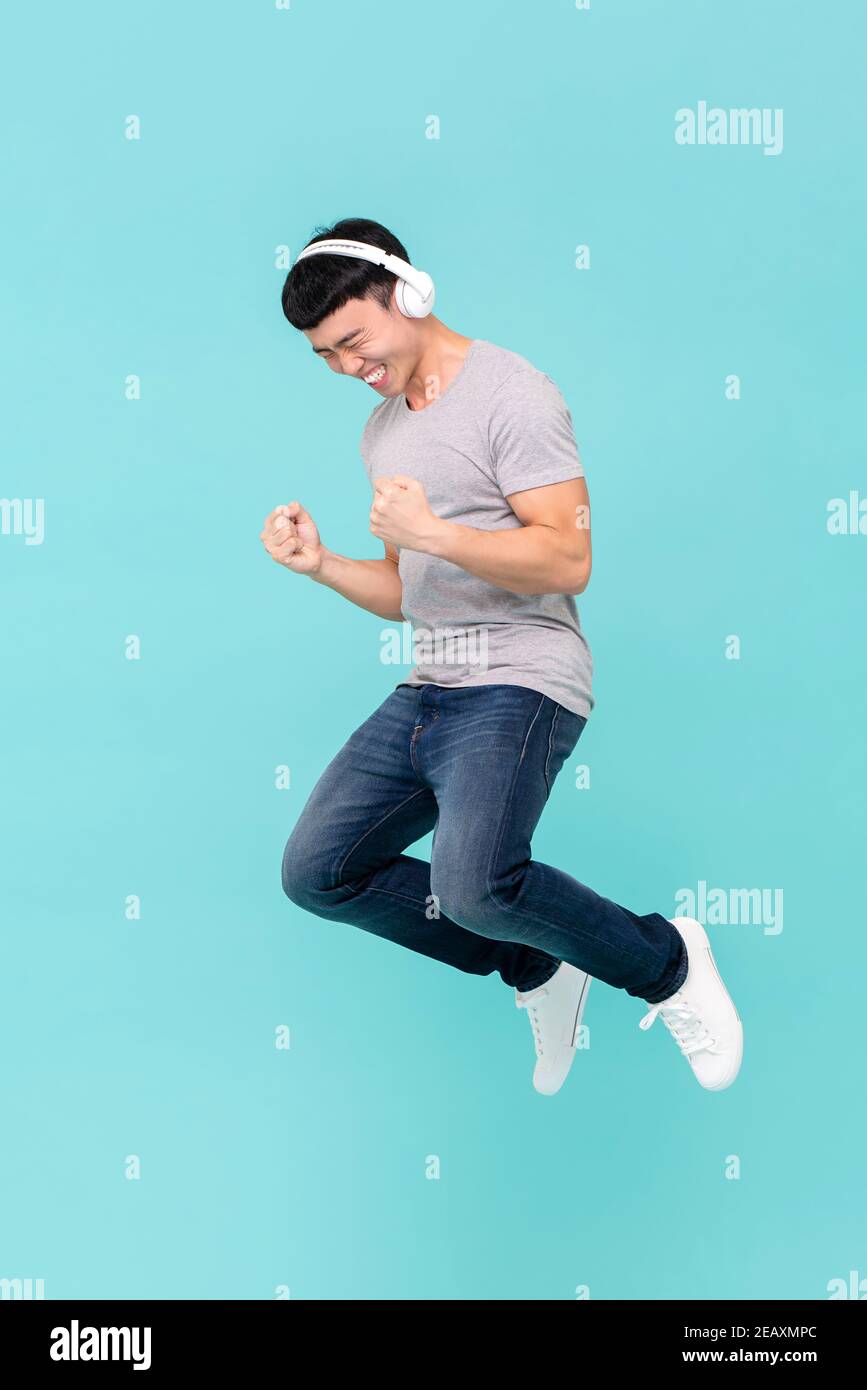 Glücklich junge asiatische Mann springen und tragen Kopfhörer hören Seine Lieblingssongs isoliert auf hellblauem Studiohintergrund Stockfoto