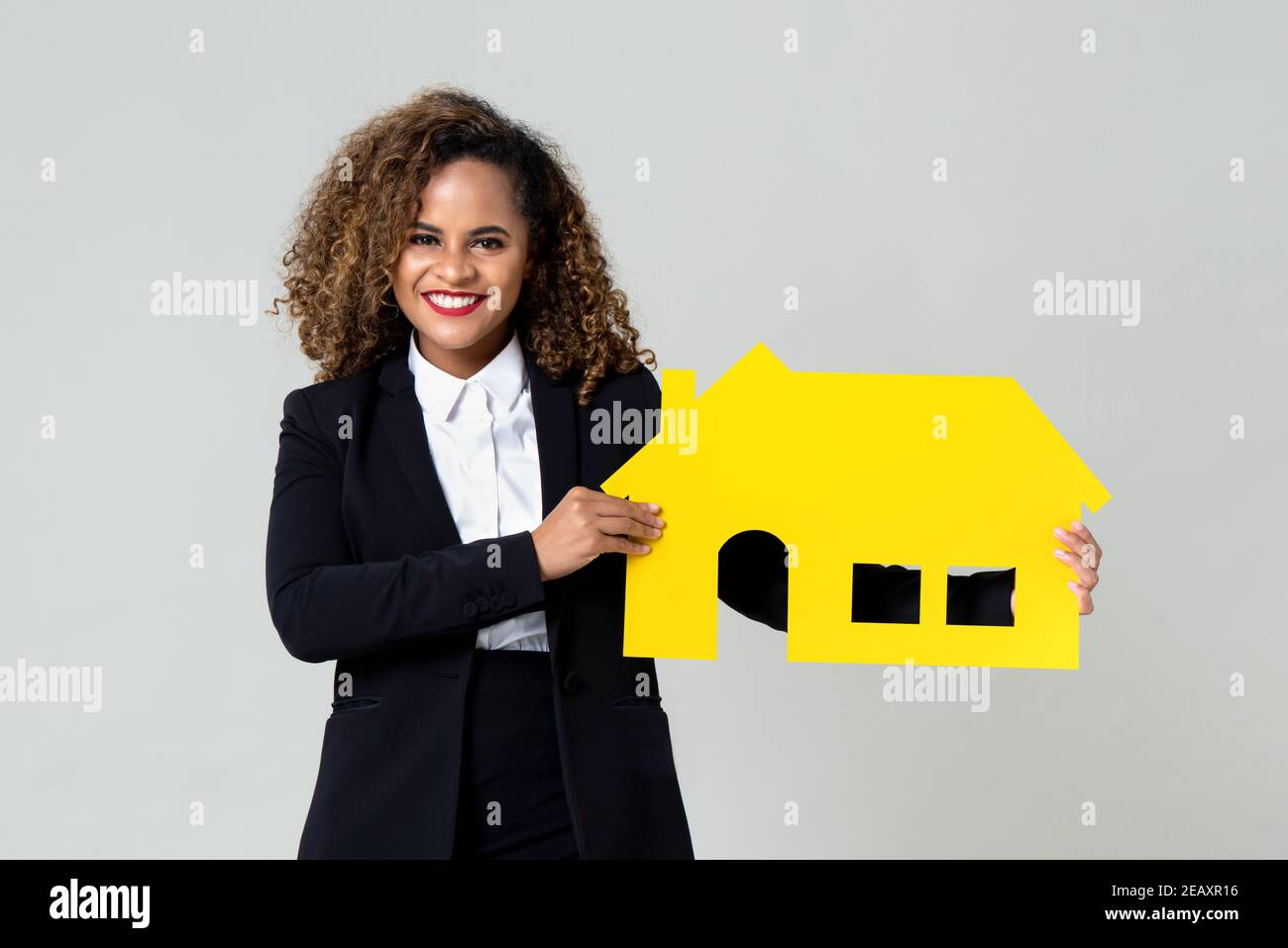 Attraktive afroamerikanische Immobilienmakler glücklich hält eine gelbe Kartongehäuse Modell isoliert auf grauem Studio-Hintergrund Stockfoto