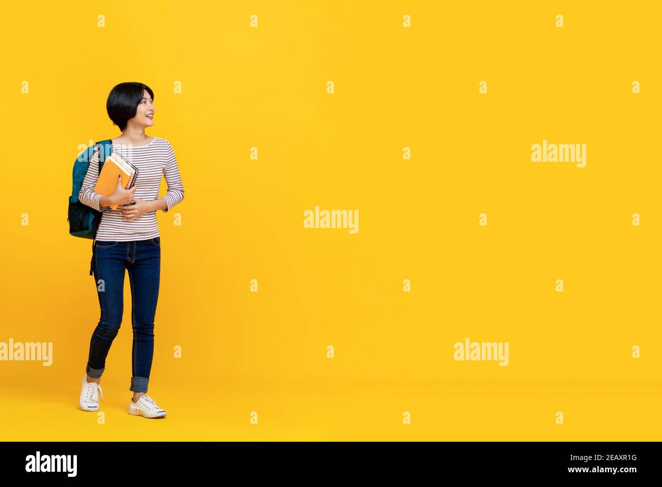 Schöne fröhliche asiatische Schüler mit Büchern und tragen Schultasche In legerer Kleidung zu Fuß weg schauen isoliert gegen gelbe Studio Hintergrund Stockfoto