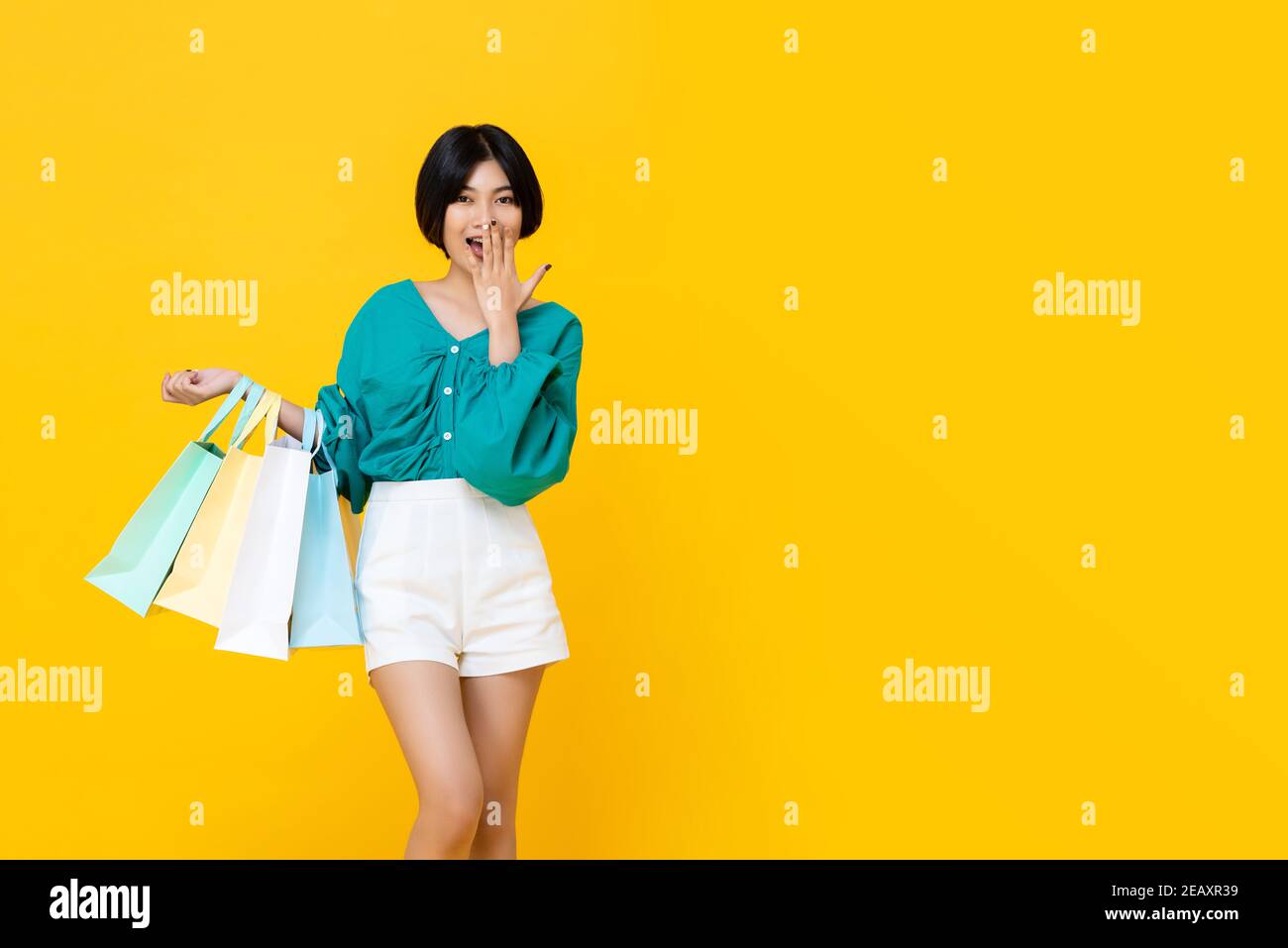 Junge fröhlich shopaholic asiatische Mädchen mit vielen Einkaufstaschen Auf ihrem Arm isoliert auf gelbem Studiohintergrund mit Kopie Platz Stockfoto