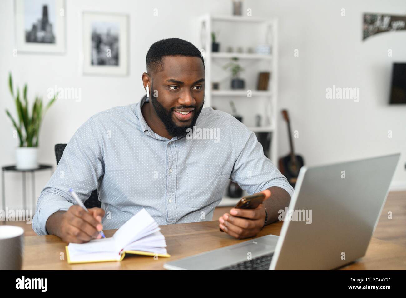 Fokussierte glückliche junge afroamerikanische Mann Student Freiberufler Notizen machen Studium mit Laptop arbeiten, tragen airpods Blick auf den Bildschirm des Stockfoto