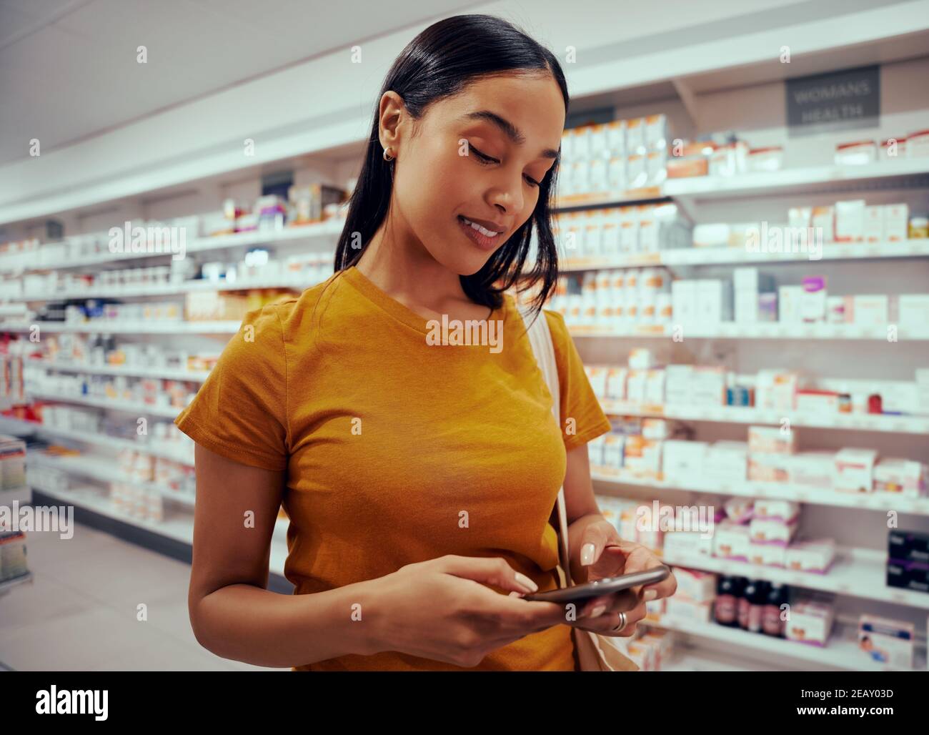 Junge Frau mit Smartphone steht gegen Regal in der Apotheke Stockfoto
