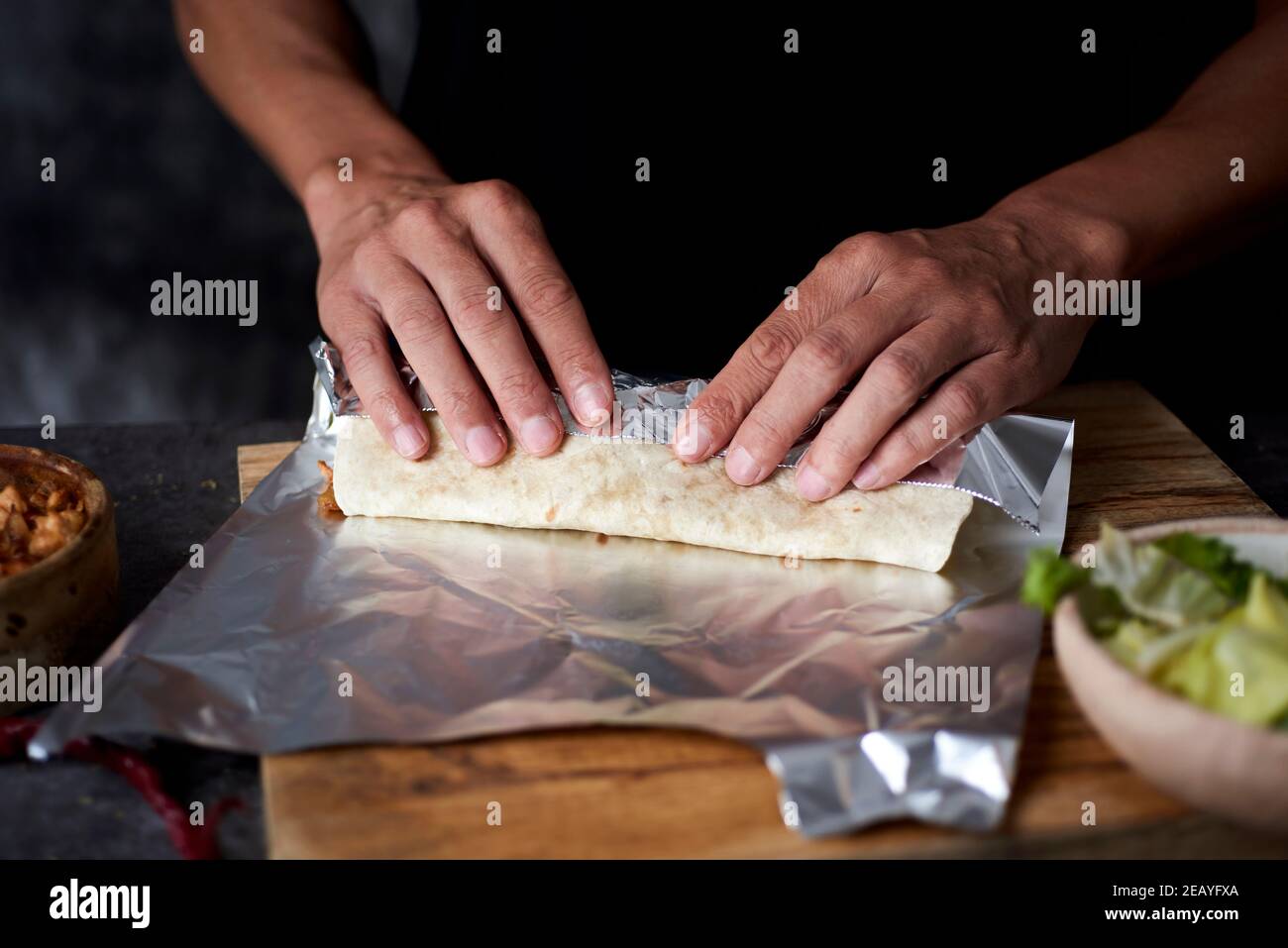 Ein junger Mann an einem Tisch wickelt in Aluminiumfolie ein Durum oder ein Burrito, gefüllt mit Hühnerfleisch mit verschiedenen Gemüse wie Zwiebeln oder rot und gekocht Stockfoto