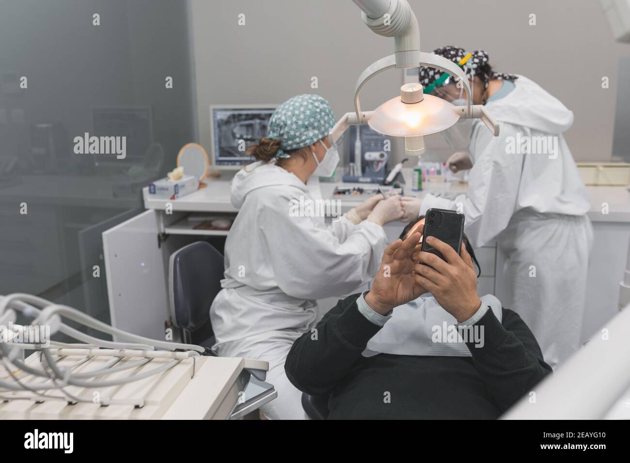 Patient mit Smartphone in der Zahnarztpraxis. Zwei Ärztinnen Vorbereitung des Verfahrens. Konzept der Zahngesundheit und Hygiene Stockfoto