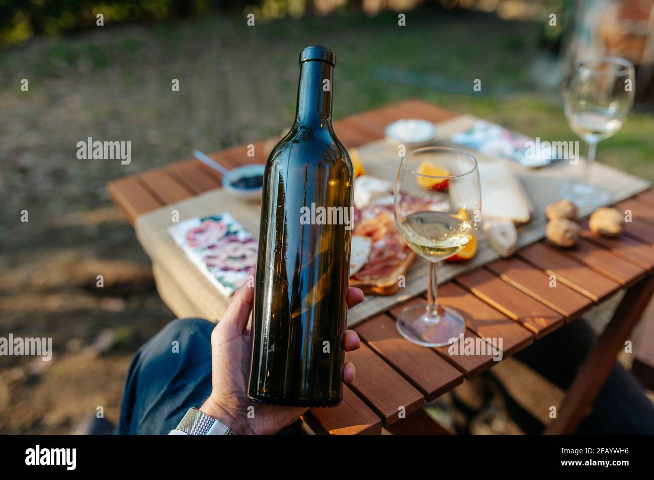 Nahaufnahme nicht erkennbare Hand hält eine Flasche Wein. Tischset mit Vorspeisen und leckeres Essen im Hintergrund. Weinprobe und Picknick-Konzept. Stockfoto