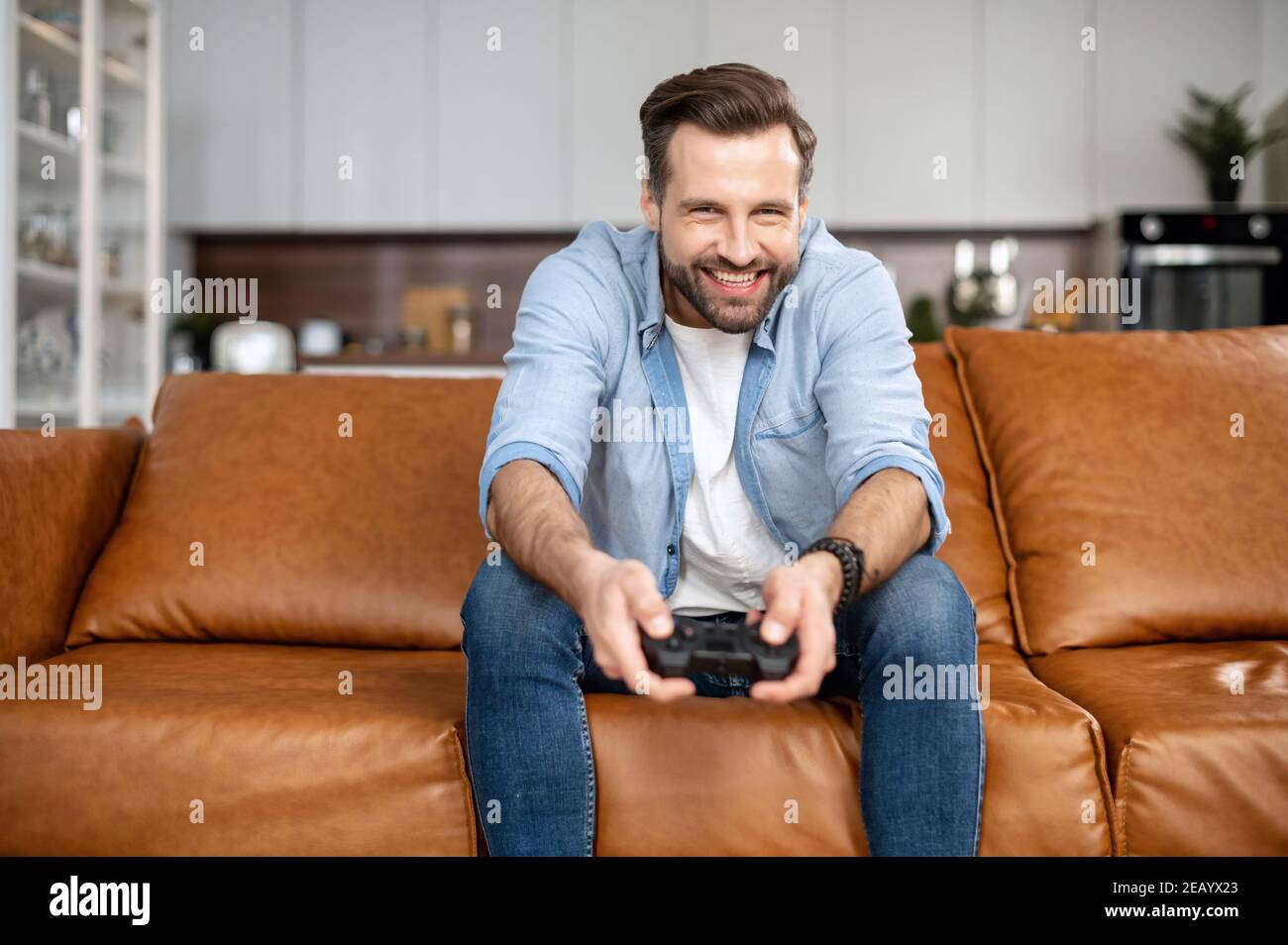Gut aussehend aufgeregt jungen Kerl verbringt Freizeit spielen Videospiel, Glücksspiel Kerl mit einem Joystick sitzt auf der Couch, spielen auf der Konsole zu Hause Stockfoto Gut aussehend aufgeregt jungen Kerl verbringt Freizeit spielen Videospiel, Glücksspiel Kerl mit einem Joystick sitzt auf der Couch, spielen auf der Konsole zu Hause Stockfoto