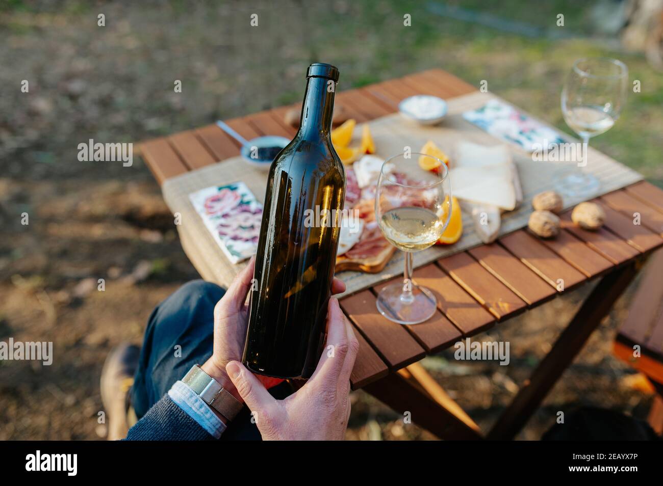 Nahaufnahme nicht erkennbare Hand hält eine Flasche Wein. Tischset mit Vorspeisen und leckeres Essen im Hintergrund. Weinprobe und Picknick-Konzept. Stockfoto
