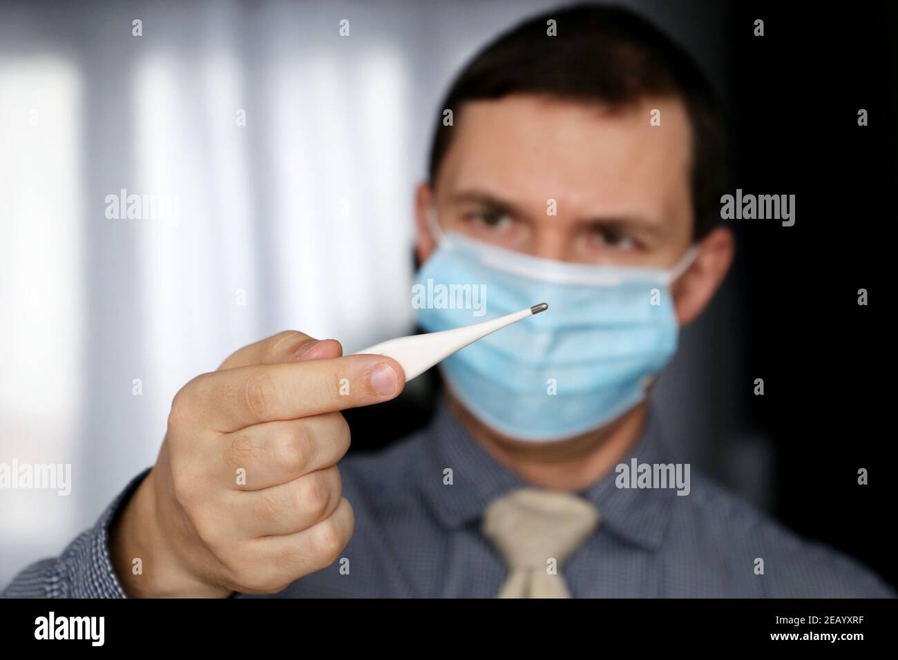 Besorgt Mann in Gesichtsmaske und Bürokleidung schaut auf Digitalthermometer in der Hand. Hohe Körpertemperatur, Sicherheit am Arbeitsplatz während einer Coronavirus-Pandemie Stockfoto