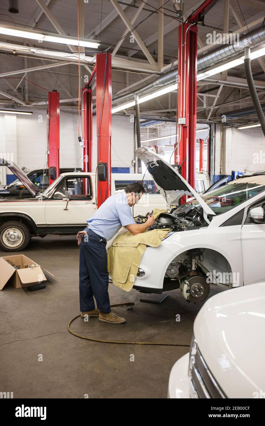 Der hispanische Mechaniker lehnt sich an ein Auto an, während er daran arbeitet Der Motorraum in einer Autowerkstatt Stockfoto