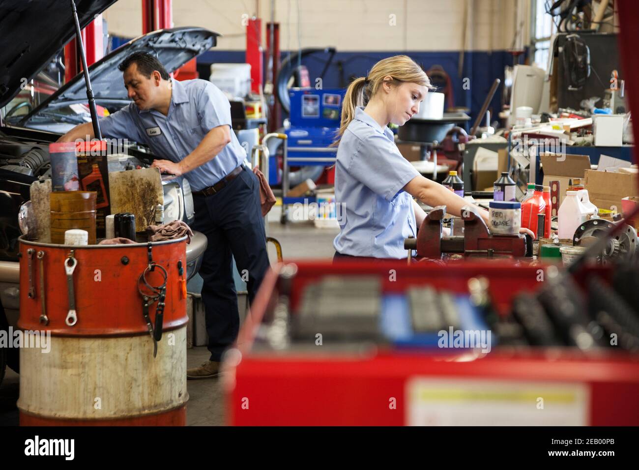 Kaukasische weibliche Mechaniker arbeitet am Motor in der Autowerkstatt Mit hispanischen Kollegen im Hintergrund Stockfoto