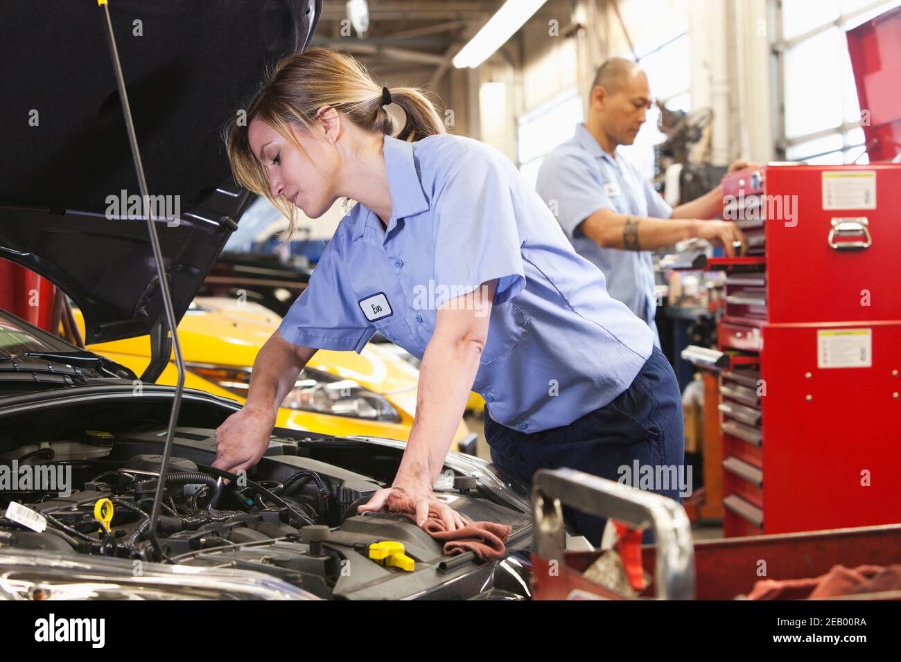 Kaukasische weibliche Mechaniker arbeitet am Motor in der Autowerkstatt Mit hispanischen Kollegen im Hintergrund Stockfoto