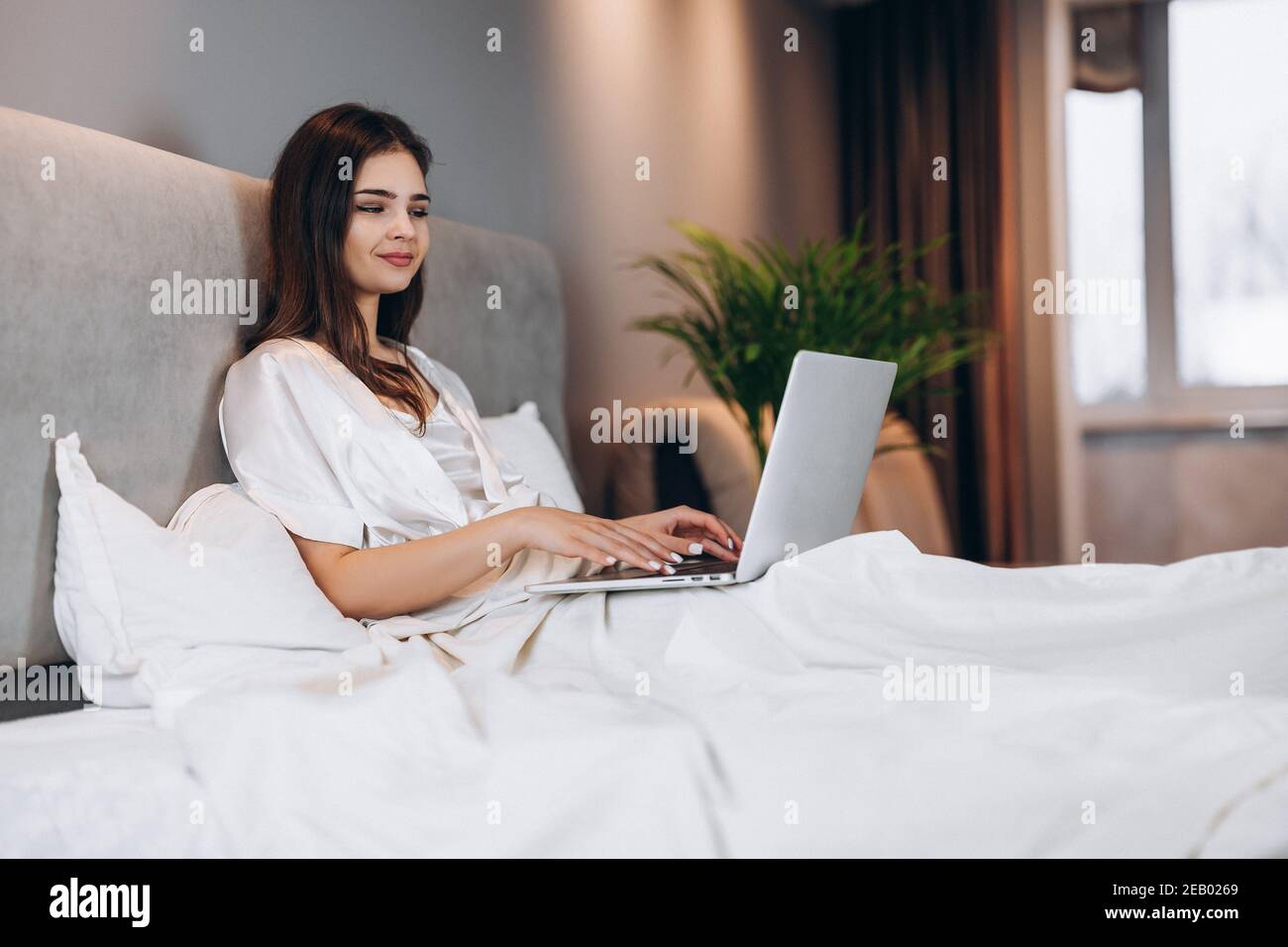 Junge schöne Frau mit Laptop im Bett. Brunette Modell mit Laptop in Seide weißen Pyjamas. Eine Frau arbeitet an einem Computer in ihrem Schlafzimmer zu Hause Stockfoto