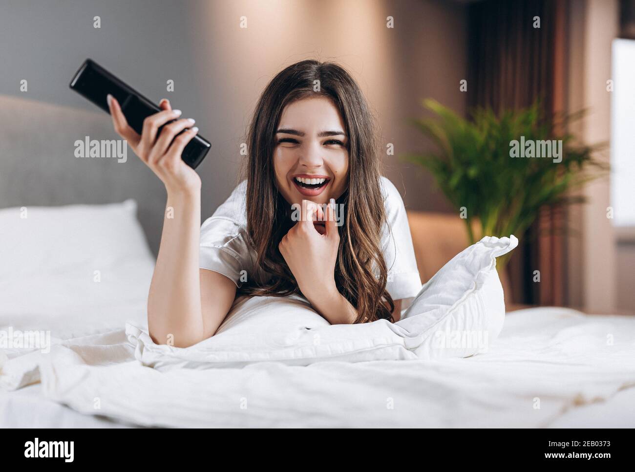 Frau in weißer Seide Pyjamas schaut TV im Schlafzimmer auf dem Bett. Weibliche Modell mit einer tv-Fernbedienung in den Händen liegend auf dem Bett. Morgen oder Stockfoto