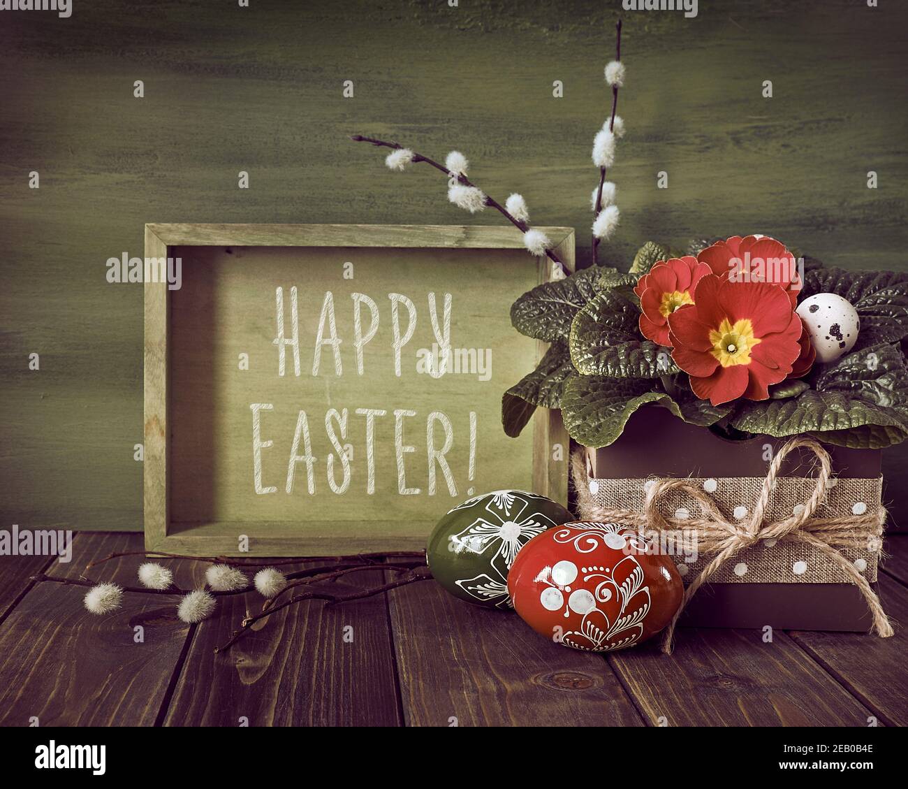 Text-Frohe Ostern. Zero Waste Ostern ohne Plastik Osterarrangement in rot, grün und erdfarben. Bemalte Holzeier. Korb mit roter Primel Stockfoto