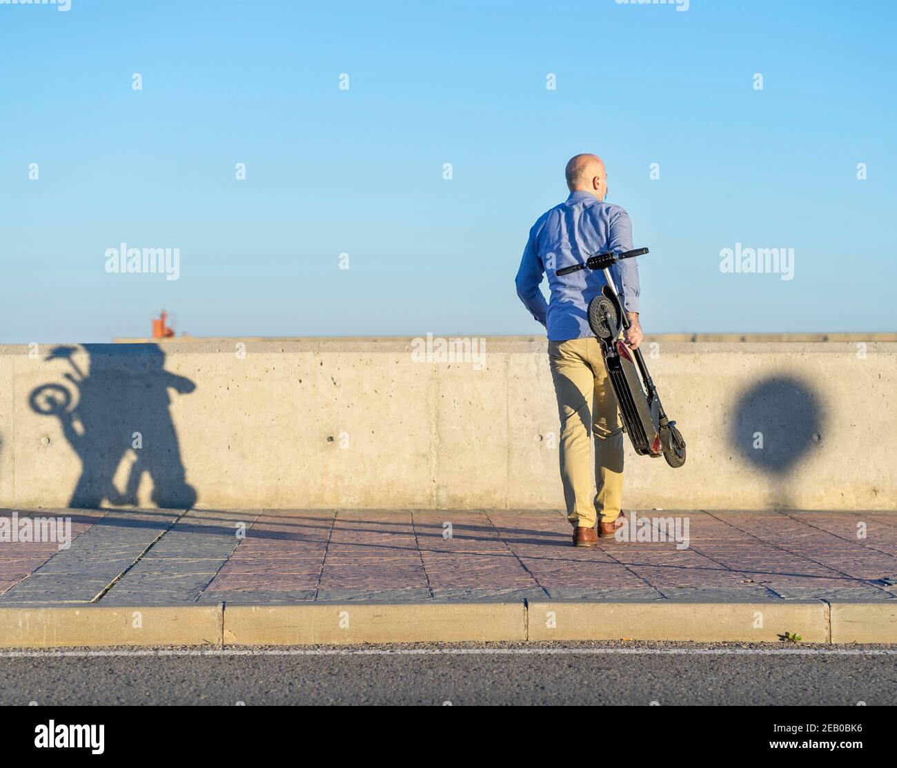 Mann mit gefaltetem Roller geht mit dem Rücken zur Kamera. Er trägt ein blaues Hemd und eine Ockerhose. Es gibt Licht bei Sonnenuntergang. Horizontales Foto Stockfoto