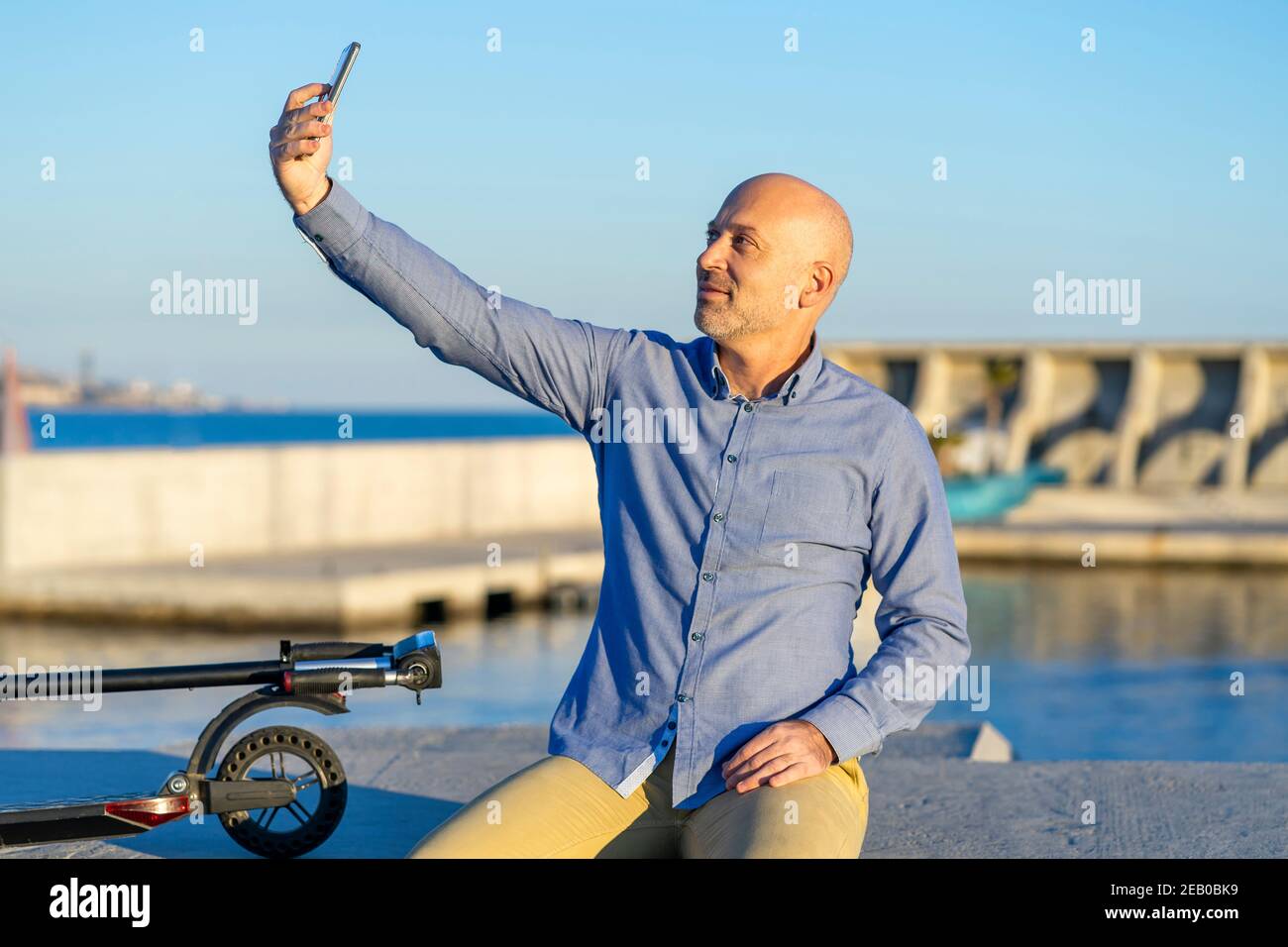 Reifer Mann nimmt Selfie mit Handy. Er sitzt und hat einen Elektroroller. Im Hintergrund sieht man das Meer. Es gibt Licht bei Sonnenuntergang. Ho Stockfoto