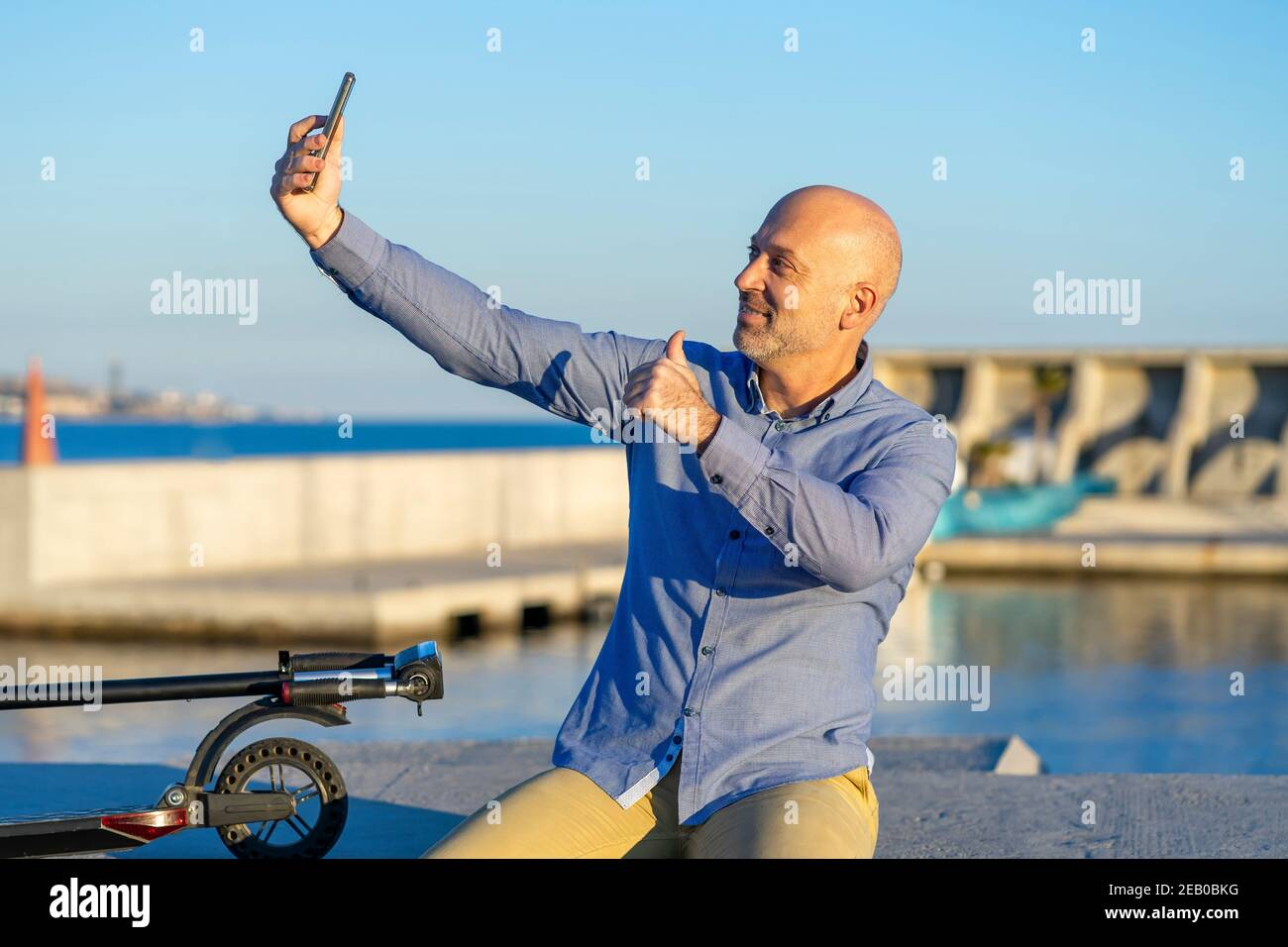 Reifer Mann nimmt Selfie mit Handy und macht Daumen nach oben Geste. Sie können das Meer im Hintergrund sehen. Abendlicht. Horizontales Foto. Stockfoto