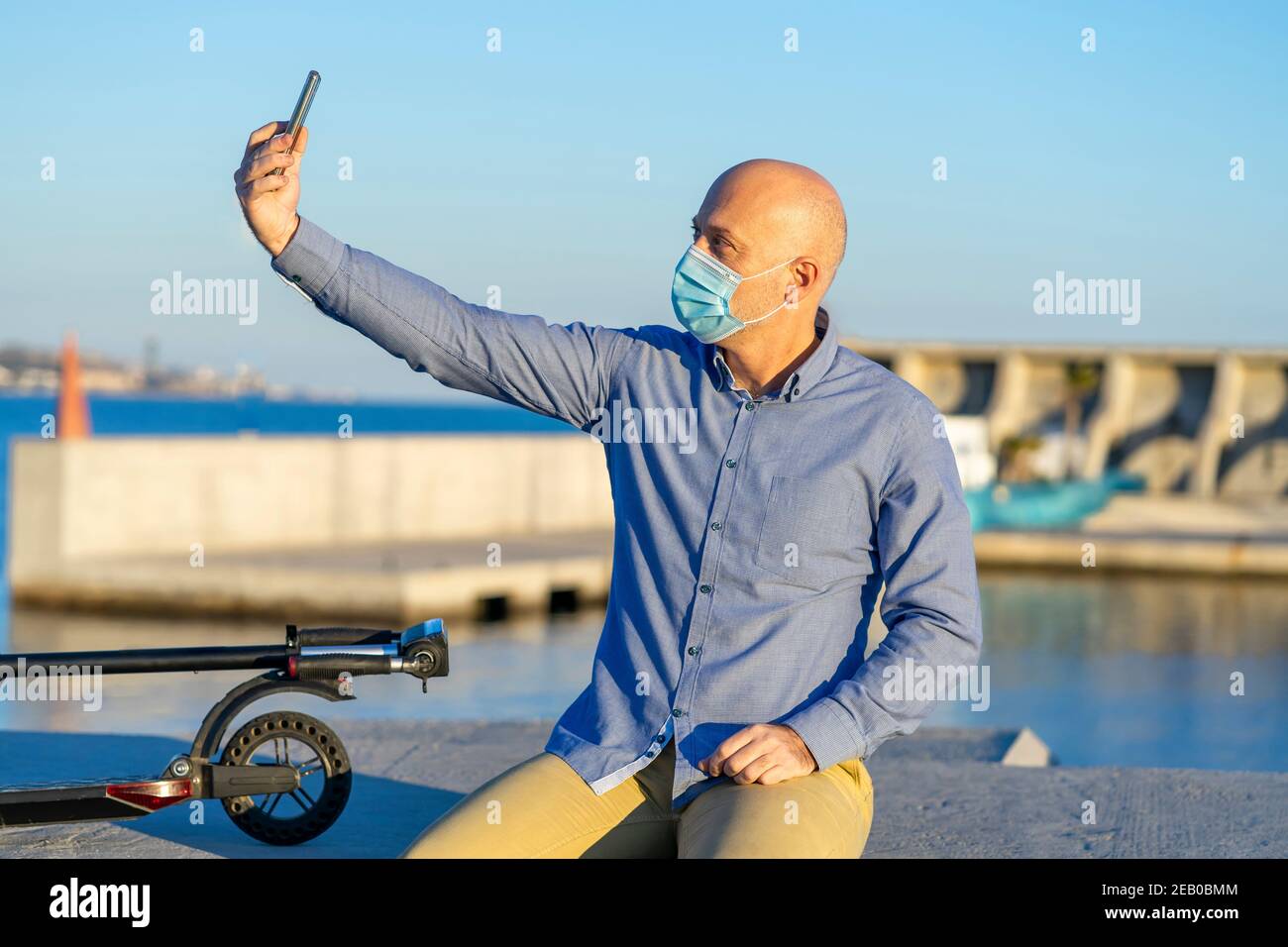 Reifer Mann nimmt ein Selfie mit seinem Handy, trägt eine Maske. Er sitzt und hat einen Elektroroller. Im Hintergrund sieht man das Meer. Stockfoto