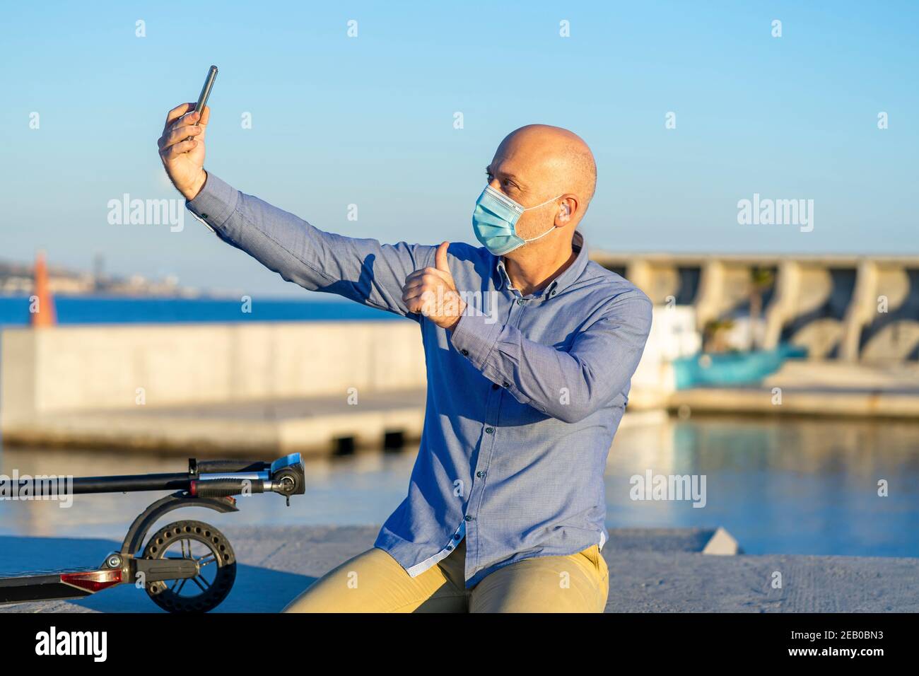 Reifer Mann nimmt Selfie mit Handy und macht Daumen nach oben Geste, trägt Maske. Sie können das Meer im Hintergrund sehen. Abendlicht. Horizontal p Stockfoto