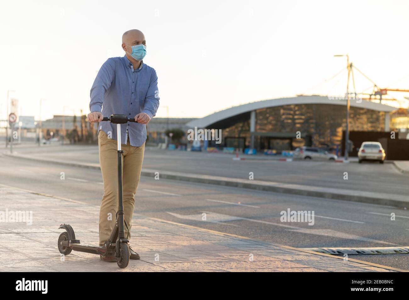 Reifer Mann trägt eine Maske nutzt einen Elektroroller in der Stadt. Er trägt blaues Hemd und ockerfarbene Hosen. Es gibt Licht bei Sonnenuntergang. Horizontales Foto Stockfoto