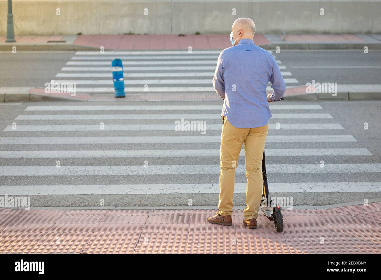 Mann mit Elektroroller wartet auf die Fußgängerüberfahrt. Er trägt ein blaues Hemd und eine gelbe Hose. Sein Rücken zur Kamera. Horizontaler Phot Stockfoto