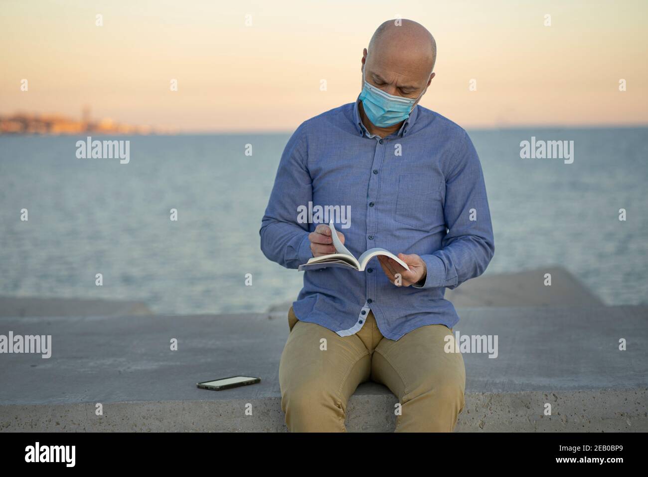 Mann liest Buch auf der Straße, trägt Maske. Das Meer ist im Hintergrund zu sehen. Er hat ein Mobiltelefon. Horizontales Foto mit Platz zum Einfügen von Text. Stockfoto