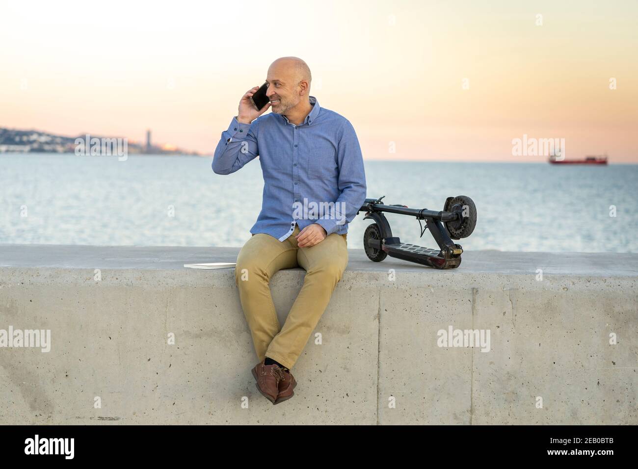 Reifer Mann spricht am Telefon. Er sitzt und mit einem Elektroroller. Er lächelt. Horizontales Foto im Freien mit dem Meer in der Backgroun aufgenommen Stockfoto