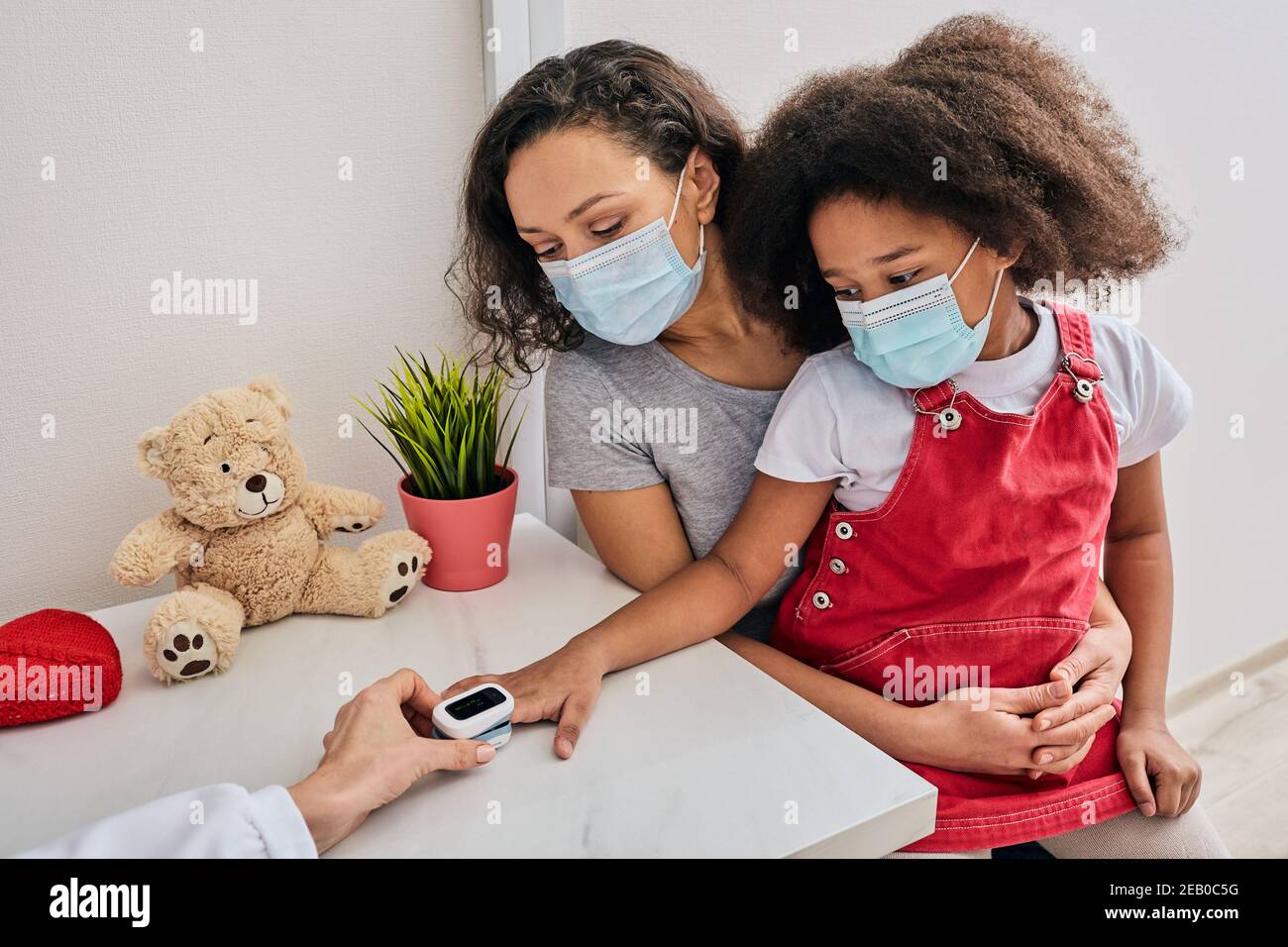 Messung der Sauerstoffsättigung und des Pulses eines Kindes. Afroamerikanische kleine Mädchen und ihre Mutter tragen medizinische Masken bei Kinderarzt Termin Stockfoto