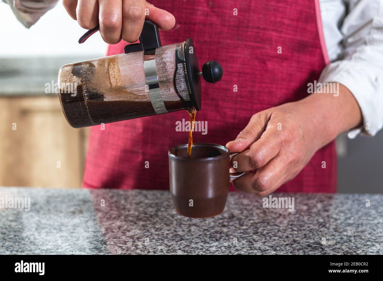 Nahaufnahme der Hände von jemandem in einer roten Schürze Gießen eine Tasse Kaffee in einer französischen Presse gebrüht. Stockfoto