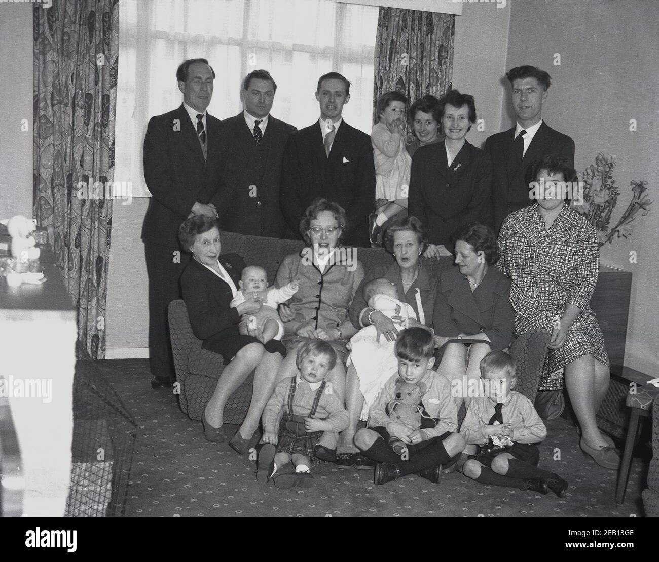 1950s, historisch, in einem vorderen Raum eines Hauses, eine große Familie versammeln sich für ein Gruppenfoto, mit vier Generationen alle zusammen, vielleicht nach der Taufe des neuen Babys, die von seiner Großmutter auf dem Sofa gehalten. Ein kleiner Junge sitzt und hält seinen Teddybär. Stockfoto