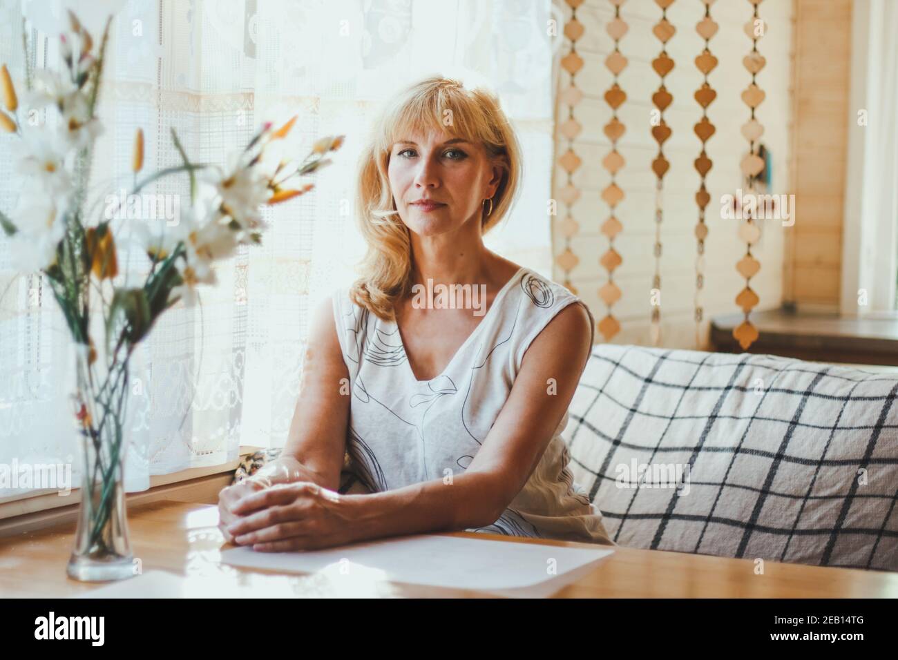 Portrait von elegant attraktive blonde Kaukasierin eine gealterte Frau mit stilvollen Haaren in weißem T-Shirt sitzt an einem Esstisch in der Nähe des Fensters, Blick auf Stockfoto