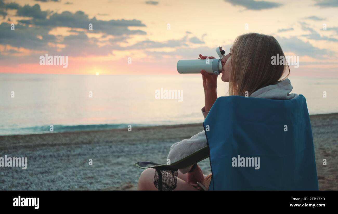 Junge Frau trinkt aus Thermoskannen und sitzt auf Campingstühlen am Strand. Profilansicht. Stockfoto