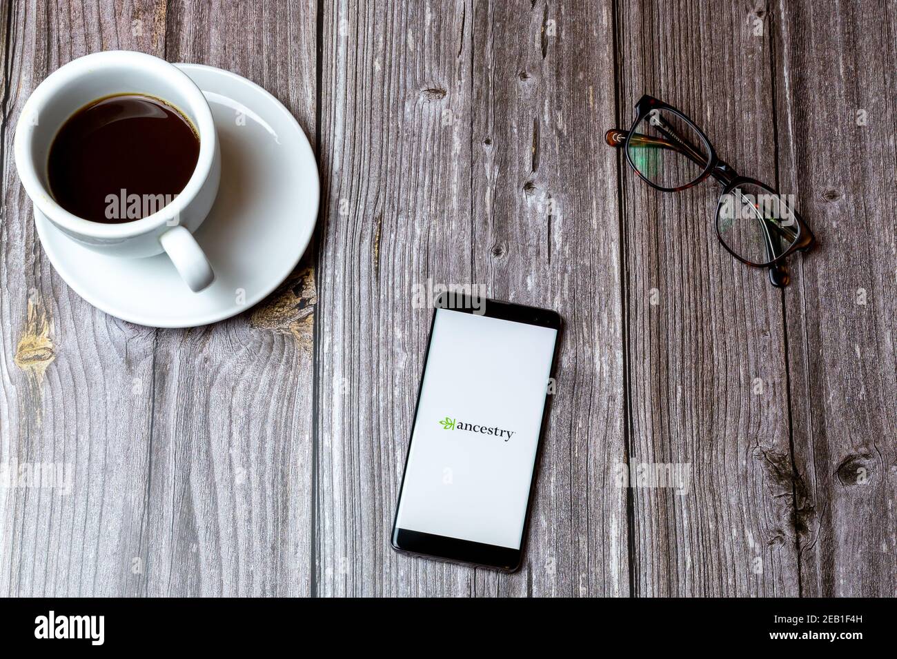 Ein Handy oder Handy auf einem Holz gelegt Tabelle mit der App Ancestry auf dem Bildschirm neben geöffnet Ein Kaffee Stockfoto