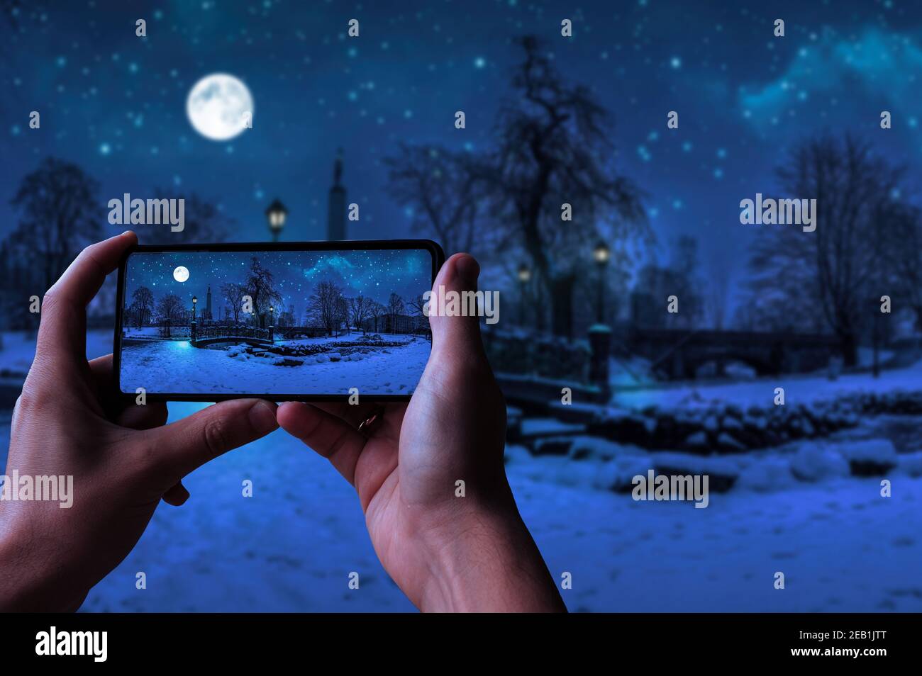 Tourist fotografieren magische Winternacht mit Sternenhimmel und Vollmond in verschneiten Park mit schöner Brücke über kleinen Teich, Straßenlicht und bedeckt Stockfoto