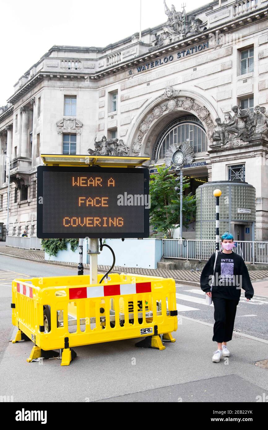 Eine Frau kommt an einem Schild mit der Aufschrift „Wear a Face Mask“ vorbei, das auf einem elektronischen Schild vor der Waterloo Station in London steht, da das obligatorische Tragen von Masken während der Pandemie Covid-19 in Großbritannien eingeführt wird. Stockfoto