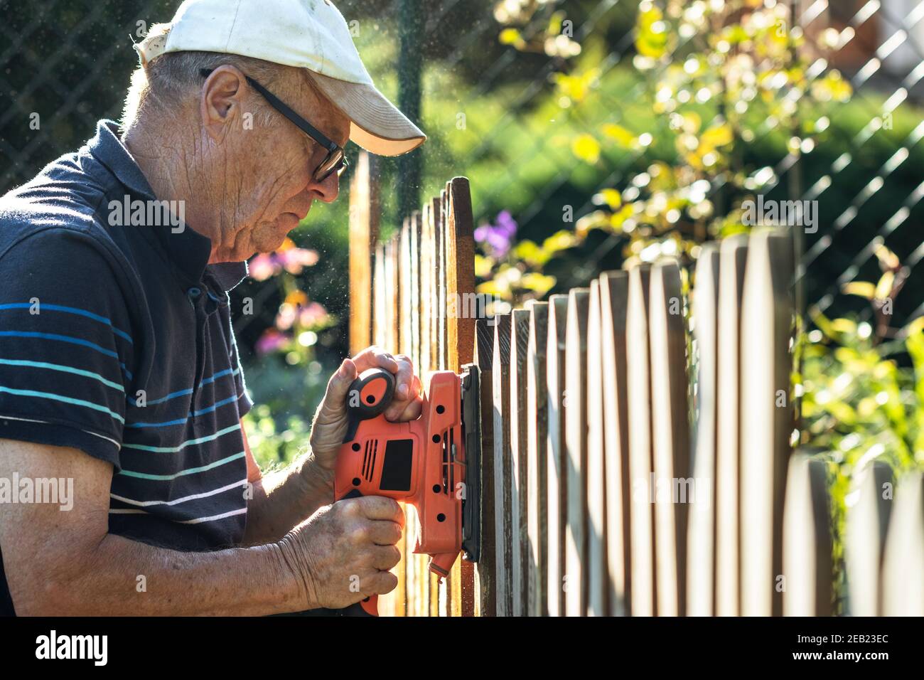 Senior Mann Schleifen Holzzaun mit elektrischem Schleifer im Garten. Reparatur Pfostenzaun im Hinterhof Stockfoto