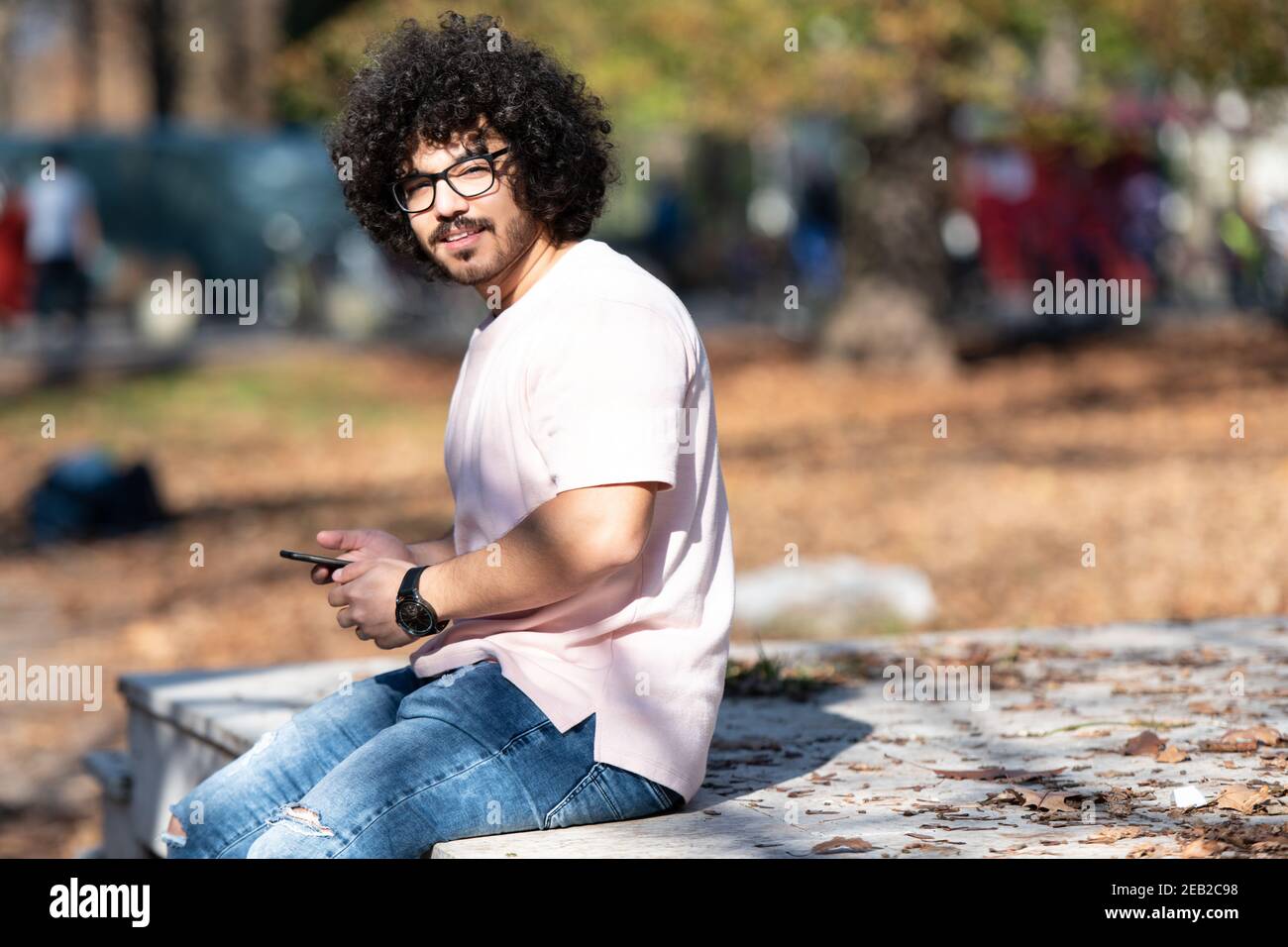 Porträt von schöner junger Afro Mann sprechen berühren Telefon oder Smartphone für den Anruf verwenden und im Herbst draußen sitzen Stockfoto