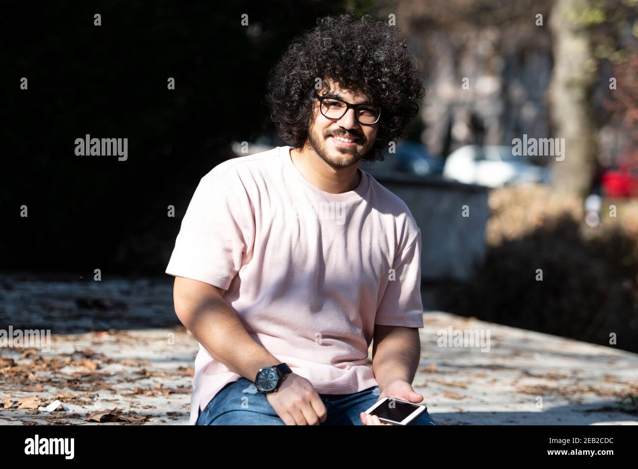 Porträt von schöner junger Afro Mann sprechen berühren Telefon oder Smartphone für den Anruf verwenden und im Herbst draußen sitzen Stockfoto