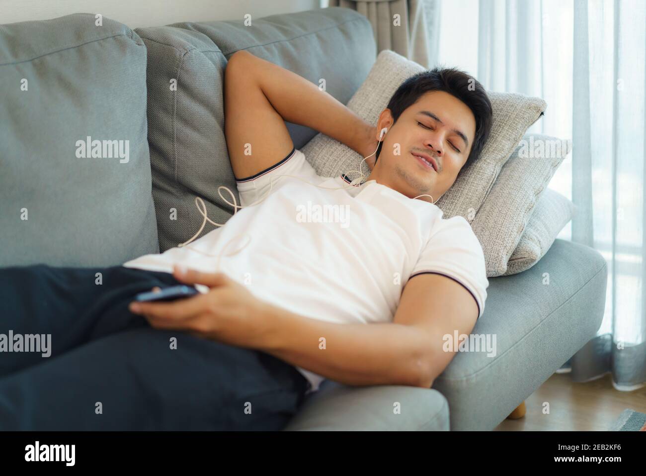 Asiatische Männer ruhen sich aus, indem sie Musik mit Kopfhörern auf dem Sofa im Wohnzimmer zu Hause hören. Stockfoto
