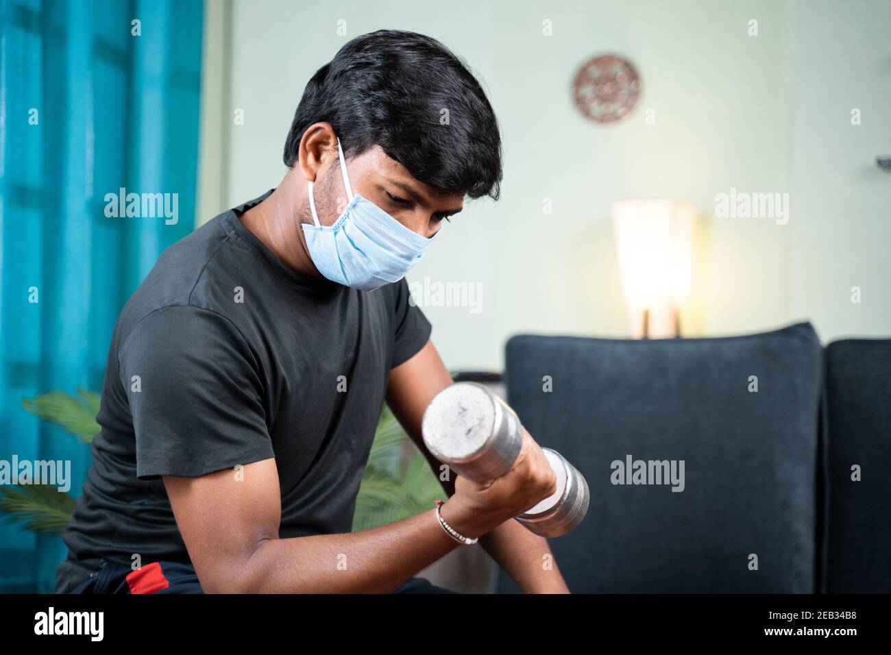 Junger Mann mit Gesichtsmaske beschäftigt in der Arbeit oder Doing Übung mit Hantel zu Hause - Konzept von zu Hause Fitness-Studio wegen Coronavirus covid-19 Pandemie Stockfoto