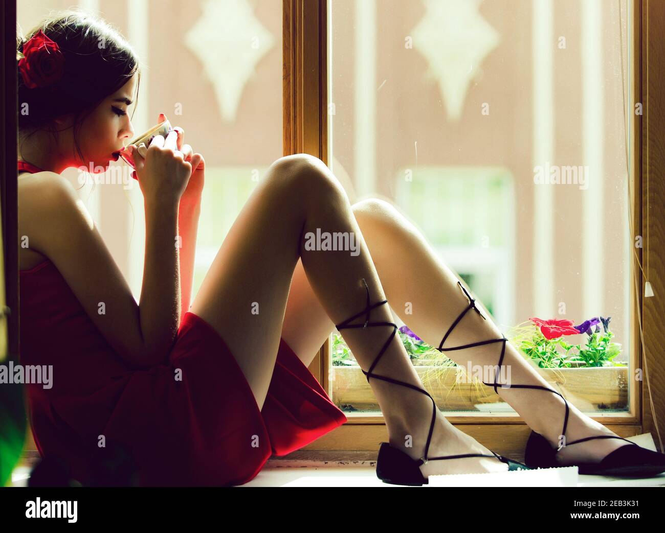 Mädchen in rotem Kleid und Rosenblüte im Haar sitzen am Fenster trinken Tee oder Kaffee aus Tasse, spanische romantische Frau denken. Stockfoto