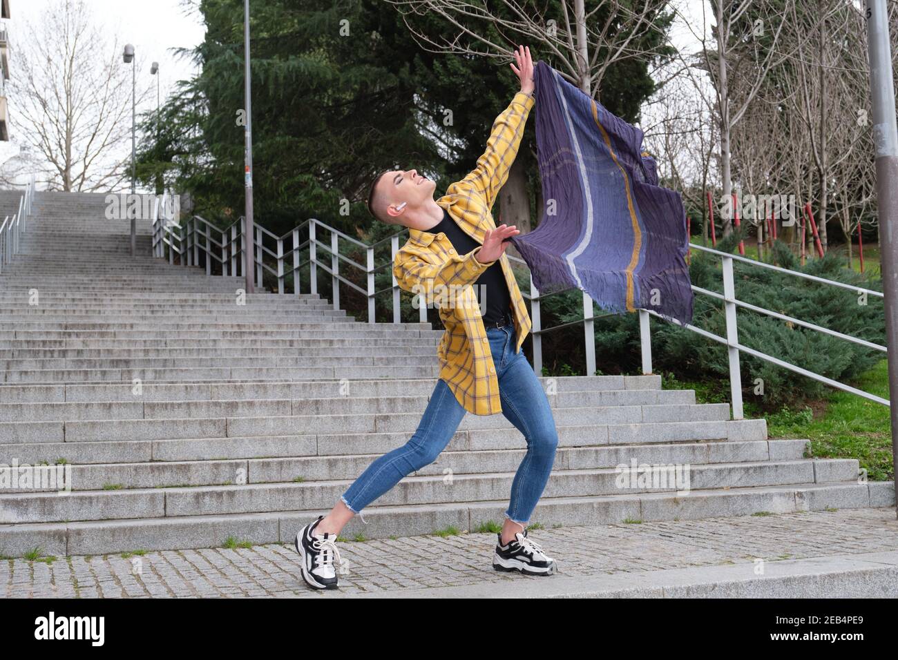 Hübscher junger Mann, der Make-up und Kopfhörer trägt, tanzt und Baumwollschal hält. Nicht binäre androgyne Kerl. Stockfoto
