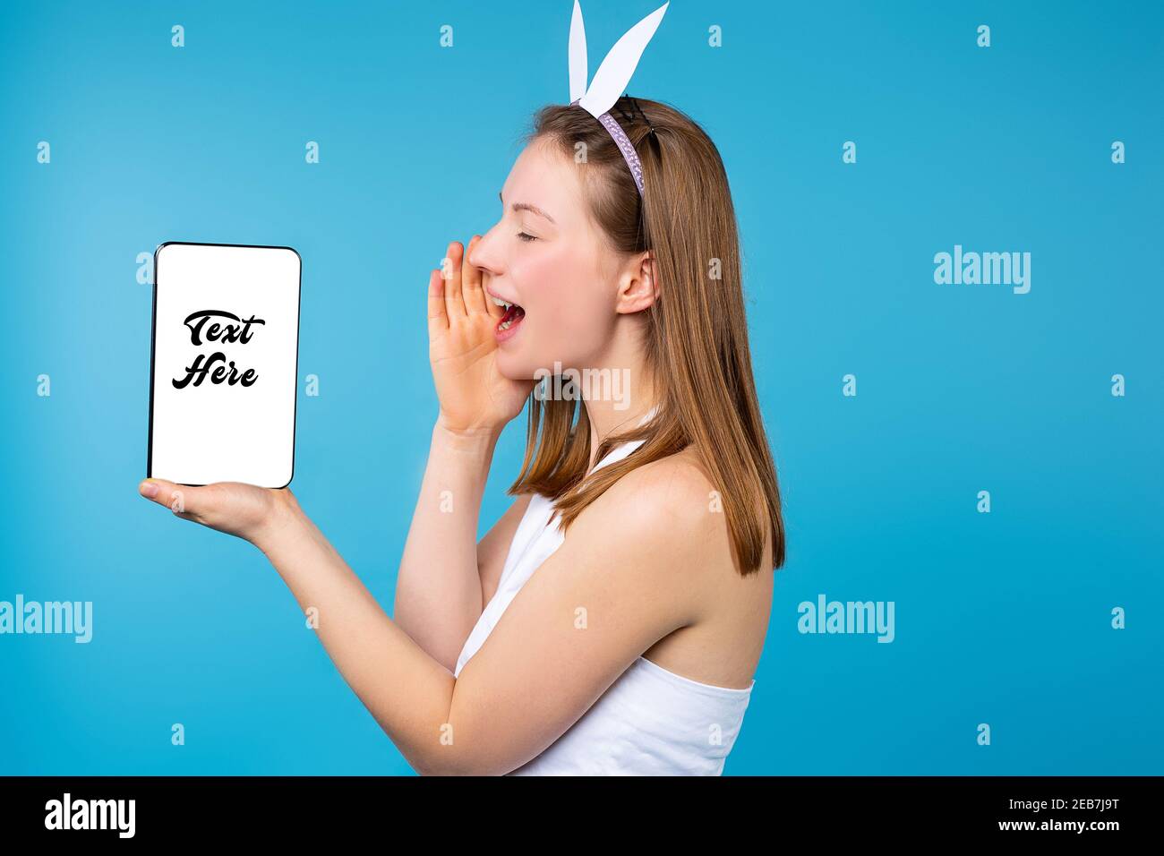 Eine Blondine mit Osterohren hält ihre Hand an ihr Gesicht und schreit auf den weißen Bildschirm der Tablette. Blauer Hintergrund. Profilfoto. Text hier eingeben. Stockfoto
