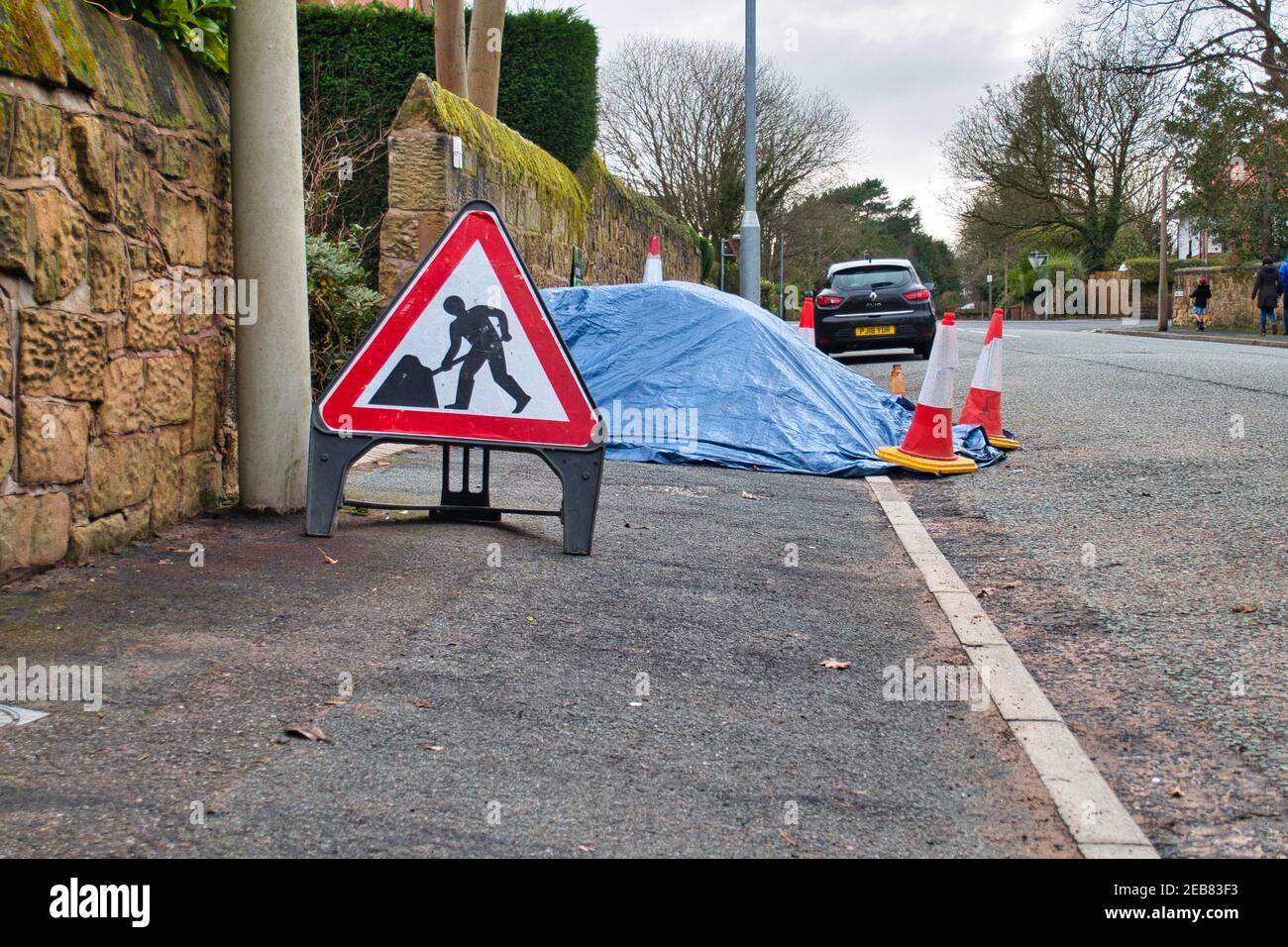 Ein dreieckiges, rot umrandtes Schild "Männer bei der Arbeit" warnt davor, dass der Straßenbelag vor dem Gebäude durch Plane überzogene Baumaterialien, umgeben von Kegeln, oder blockiert ist Stockfoto