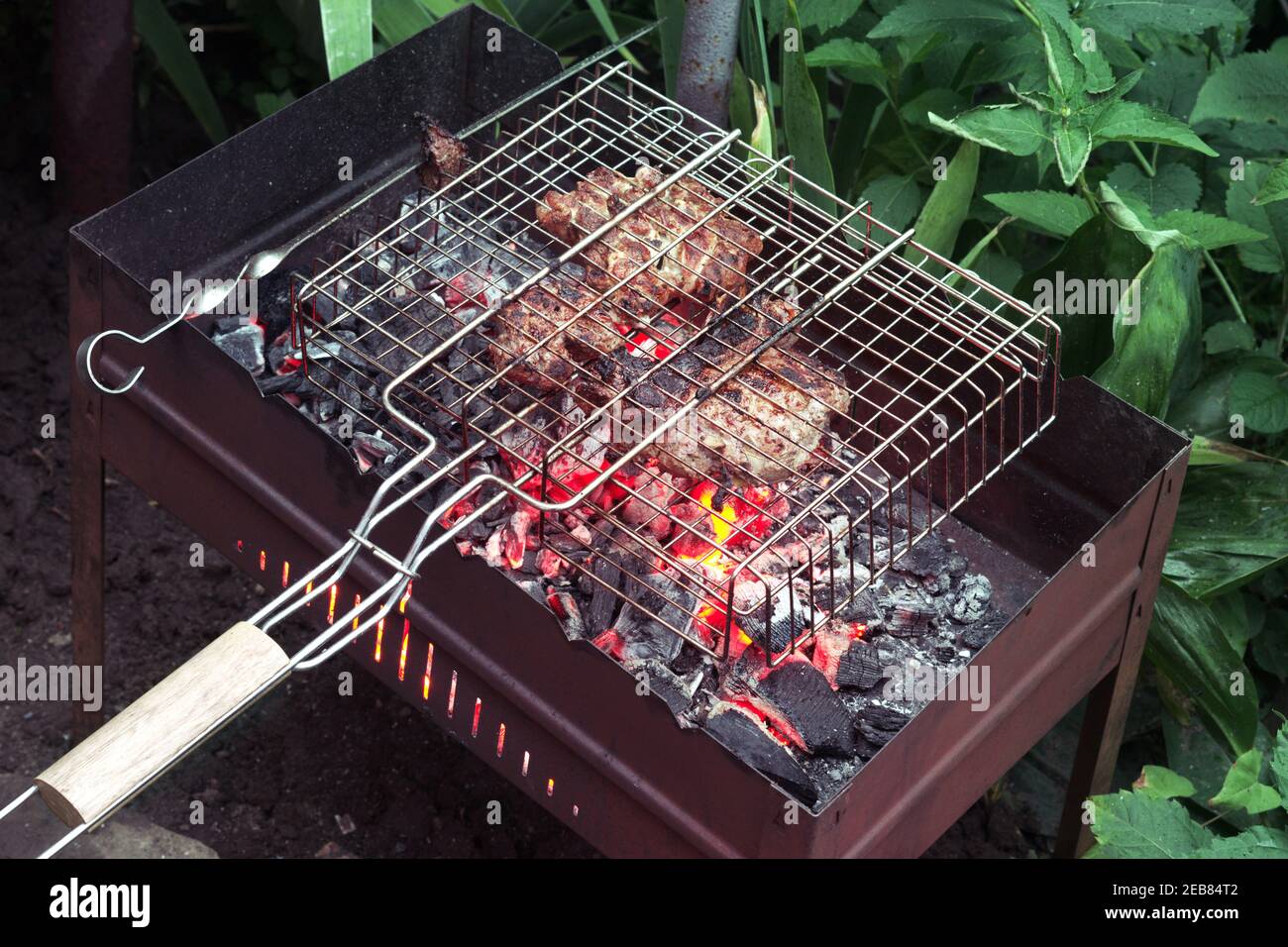 Mariniertes Fleisch im Grillkorb und auf flachem Stahlspieß, gekocht auf rot-heißer Holzkohle in einem Holzkohlegrill. Auf dem Boden unter dickem grünem Gras installiert Stockfoto